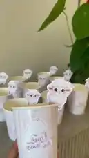 A charming display of personalized paper cups decorated with cute lamb cutouts and Arabic text reading 'عيد السعيد تفويه' (Happy Eid Al-Adha). These festive party supplies, perfect for celebrating Eid Al-Adha, feature elegant white and pink designs with pearl embellishments. The scene captures the joyful spirit of the holiday with these delightful decorations. by @rubaaomaar with Original Sound by rubaaomaar