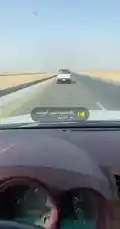 This POV footage captures a journey on a wide, multi-lane highway cutting through a vast desert landscape. The camera follows a white Toyota pickup truck ahead, showcasing the open road under a clear sky. Arabic text overlays suggest a promotional message for 'TASI digital platform,' possibly related to vehicle services or logistics. by @s3432x with Original Sound by s3432x
