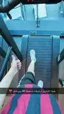 A young man in a red and black striped shirt and black shorts performs a series of arm stretches and warm-up exercises in a modern gym with red and black decor. The setting includes various weight machines and mirrors. The video then transitions to a first-person view of someone running on a treadmill, showcasing a panoramic city skyline through large windows. Text overlays in Arabic provide context about the workout routine. by @by.sul with by.sul by Original Sound