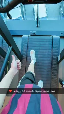 A young man in a red and black striped shirt and black shorts performs a series of arm stretches and warm-up exercises in a modern gym with red and black decor. The setting includes various weight machines and mirrors. The video then transitions to a first-person view of someone running on a treadmill, showcasing a panoramic city skyline through large windows. Text overlays in Arabic provide context about the workout routine. by @by.sul with by.sul by Original Sound