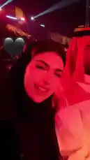 A woman with dark hair and striking red lipstick, wearing a black hijab, smiles and poses for a selfie in a vibrant red-lit environment. She points towards her companion, a man dressed in traditional Saudi attire including a white thobe and a red-and-white patterned ghutra. The background suggests they are at a lively event, possibly a concert or festival, with dramatic stage lighting and a crowd visible. A heart emoji is overlaid on the screen, adding a personal touch. by @soja_design with Original Sound by soja_design