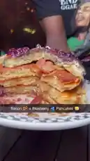 A close-up shot reveals a towering stack of golden-brown pancakes, generously filled with crispy bacon and drizzled with rich, dark berry compote. A hand enters the frame, pouring warm maple syrup over the top, causing it to cascade down the sides and pool on the plate. The focus is entirely on the delicious details of the food, highlighting its texture and appetizing appearance. by @marvedtv with Jimmy Cooks by Drake, 21 Savage