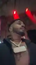 JUSTIN MARC is captured in a candid, late-night selfie-style vlog walking through a city street. He speaks directly to the camera while showcasing his signature style, including a large diamond-encrusted chain with a rectangular pendant and visible tattoos on his chest and neck. The urban backdrop features brick buildings illuminated by striking red lights, creating a moody atmosphere. by @sexxyjustin with Original Sound by sexxyjustin