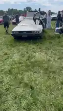 This video captures a silver DeLorean DMC-12, famous for its role in 'Back to the Future,' parked on a grassy field at what appears to be a car show. The iconic gull-wing doors are open, showcasing the vehicle's unique design to onlookers under a partly cloudy sky. by @tershine with Original Sound by tershine