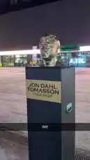 A nighttime view of a bronze bust statue dedicated to Jon Dahl Tomasson, displayed on a pedestal with his name and years '2024-2025'. The camera pans around the sculpture, highlighting its reflective surface against the illuminated backdrop of the Strawberry Arena. by @fabsvo with Original Sound by fabsvo