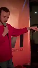 A man in a red sweater performs stand-up comedy at a club, asking the audience for drink suggestions. He playfully calls out 'David' before suggesting 'Copacabana' as a name for a mixed drink, creating a lighthearted moment. This scene captures the essence of live comedy and audience interaction. av @sommarkattens med Original Sound av sommarkattens