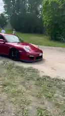 A red Porsche 911 Turbo S is seen drifting on a dirt road in a scenic outdoor setting. The car performs a controlled slide, showcasing its power and handling. A person is briefly visible near the rear of the vehicle. The surrounding area features grassy fields and trees under a partly cloudy sky. by @tershine with Original Sound by tershine