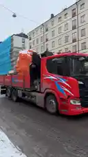 A massive, oversized orange head sculpture is being transported through city streets on a red flatbed truck belonging to 'Haninge Åkeri'. The truck, which advertises 'Biogas!' on its side, navigates an urban intersection past apartment buildings under an overcast sky. Pedestrians, including one pushing a stroller, pause to watch the unusual spectacle pass by. by @fabsvo with Original Sound by fabsvo