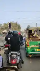 This scene shows a busy road intersection in India where a train passes overhead. Traffic, including a green auto-rickshaw and several scooters with riders wearing helmets, is stopped at a red signal. The perspective is from behind the vehicles, giving a realistic view of daily commuting challenges near railway lines. by @guptaji_47 with guptaji_47 by Original Sound