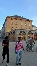 In this vlog-style clip, content creator IZER walks through a charming European city square, showcasing his streetwear look. He films himself in a black Nike t-shirt, gold chain, and backwards cap while interacting with a friend. They stroll past historic buildings and local shops, including a bookstore named 'Poésie,' capturing the relaxed atmosphere of an afternoon in Europe. by @izerflyy with Original Sound by izerflyy