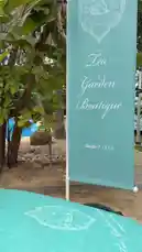 This video showcases a beautifully arranged outdoor tea party setup at 'The Garden Boutique,' featuring elegant tiered trays with pastries, sandwiches, and coffee, all set against a stunning tropical backdrop with a swimming pool and lush greenery. A staff member is seen serving drinks, while the tour continues along a stone path through a serene garden leading to a rustic building, capturing the essence of a relaxing resort experience. by @nowaira-11 with original sound
