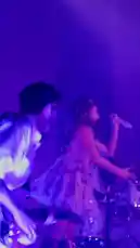 Witness EMMA 🎀 delivering an energetic live performance on a vibrant stage. Dressed in a charming pink mini dress adorned with bows and striking red boots, she sings passionately into a microphone, captivating the audience. The concert features dynamic lighting effects, with dramatic spotlights and colorful backdrops shifting between purple, blue, and red hues. Band members, including a keyboardist and guitarist, accompany her on stage. The crowd is visibly engaged, many recording the show on their smartphones, creating a lively atmosphere filled with music and excitement. by @emmaaamusic with emmaaamusic by Original Sound