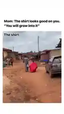 In a heartwarming and humorous scene set in a rural village, a young child walks down a dusty path wearing a massive red shirt that clearly belongs to their mother. The shirt is comically oversized, making the child look like they're wearing a cape or a small tent. An adult, presumably the mother, walks beside them. Text overlays on the screen read 'Mom: The shirt looks good on you. "You will grow into it"' and 'The shirt:', adding a layer of comedic irony to the situation. The child continues walking, showcasing the absurdly large garment, while other villagers and animals move about in the background, creating a candid and endearing moment. by @levis_ian with Original Sound by levis_ian
