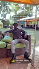 A man, identified as Gxcci Outta Here, is seen relaxing on a wicker sofa under a large orange umbrella at the Sheraton Kampala Hotel. He is dressed in a patterned shirt, light-colored jeans, and black boots. The setting is a lush, green outdoor patio area with other guests visible in the background. The scene is accompanied by a hip-hop track with lyrics about travel, wealth, and past experiences. by @managergxcci with can't leave without it by 21 Savage