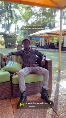 A man, identified as Gxcci Outta Here, is seen relaxing on a wicker sofa under a large orange umbrella at the Sheraton Kampala Hotel. He is dressed in a patterned shirt, light-colored jeans, and black boots. The setting is a lush, green outdoor patio area with other guests visible in the background. The scene is accompanied by a hip-hop track with lyrics about travel, wealth, and past experiences. by @managergxcci with can't leave without it by 21 Savage