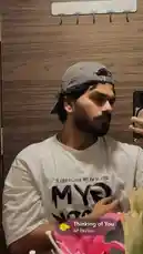 This static mirror selfie features D SUN 🇦🇪🇮🇳 in a casual moment. He wears a backwards grey baseball cap and a white mesh t-shirt with 'BELIEVE IN YOURSELF MY' printed on it. In his hand is a smartphone, suggesting he is recording a personal clip. A prominent music overlay displays the song 'Thinking of You' by AP Dhillon, indicating a relaxed, reflective mood. The setting appears to be a bathroom with wood-paneled walls and a white hook rack above. by @dsunmusic with Thinking of You by AP Dhillon