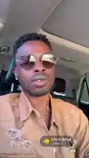 In this short clip, musician Delly is seen sitting in the driver's seat of a car, looking directly at the camera. He is wearing aviator sunglasses and a tan, distressed button-up shirt. A music player overlay indicates the song 'Life Is Good' by Lil Man J is playing, setting a positive and relaxed tone. by @delwinbazilme with Life Is Good by Lil Man J