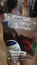 A close-up shot shows a hand holding a chilled bottle of Michelob Ultra beer next to a bag of Takis Chismes spicy snacks. The setting appears to be inside the Corpus Christi Trade Center in Texas, with people visible in the background. A regional Mexican song plays in the audio, creating a casual, social atmosphere. by @tomasg3901 with Los Chismes by Chalino Sanchez, Los Amables Del Norte