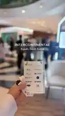 A close-up view from designer Abdullah's perspective at the InterContinental hotel in Riyadh, Saudi Arabia. The shot focuses on a hand holding a conference badge with sponsor logos, set against the bustling, elegant backdrop of the event space. by @abood114 with الدخول العظيم by عبود