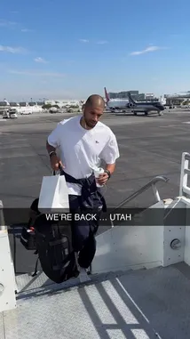 A behind-the-scenes moment shows an LA Clippers player arriving at the airport, carrying his gym bag and coffee. He walks down the aircraft stairs under a clear blue sky, dressed casually in a white t-shirt and navy pants, giving fans a glimpse into the daily routine of a professional athlete. by @clippers with Original Sound by clippers