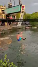 This short clip captures a man leaping from a suspended platform into a body of water. The platform, which features a banner for 'Soccer Land,' is lifted by a crane. The man executes a dive, hitting the water with a large splash. Other people are visible in the water nearby, and the background shows a beach area with spectators under umbrellas. by @digiflips with Original Sound by digiflips