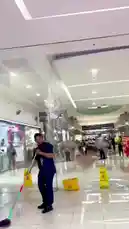 A dramatic water leak occurs inside a shopping mall, causing a large volume of water to pour down from the ceiling onto the tiled floor. A janitor in blue work clothes rushes to clean up the mess using a mop and bucket while other shoppers walk past the flooded area. The incident creates a hazardous situation requiring immediate attention. by @eirakoze64 with Original Sound by hadiakhan7689