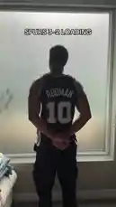 A man identified as Jeremiah Brown stands with his back to the camera, wearing a black San Antonio Spurs jersey with 'RODMAN 10' on the back. He is looking out a large window, seemingly watching a game. Text overlay reads 'SPURS 3-2 LOADING'. A male voice says, 'Yo. It's time.' The man then turns around and walks away. by @findjeremiah with Original Sound by findjeremiah