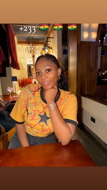 In a warm, intimate setting, Everything Naddy sits at a wooden table, looking directly into the camera with a soft smile. She wears a vibrant yellow graphic t-shirt featuring a prominent star design. Her hair is styled back, and she accessorizes with a chunky silver bracelet. The background reveals an elegant interior with chandeliers and ambient lighting, suggesting a restaurant or lounge. As she rests her chin on her hand, her expression is thoughtful and engaging, creating a personal connection with the viewer. by @everythingnaddy with I Love you ❤️ by Mildred