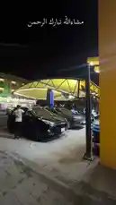 This nighttime scene captures the bustling atmosphere of 'مغسلة أربع قطرات' car wash in Qatar. Under a large yellow canopy illuminated against the dark sky, several cars—including a black Hyundai—are being cleaned by staff using hoses and cloths. The setting suggests a professional, well-lit outdoor service area where customers can get their vehicles washed after hours. by @wash.4drops with Original Sound by wash.4drops