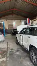 This video offers a behind-the-scenes look inside the Wasta Hawkar service center, showcasing a professional automotive workshop environment. Viewers can see vehicles like a white SUV and a black car undergoing maintenance, with a blue hydraulic lift visible in the background. The setting features a high ceiling, industrial lighting, and prominent branding for 'WASTA HAWKAR SERVICE', providing insight into the daily operations of a modern auto repair facility. by @hawkar_service with Original Sound by hawkar_service