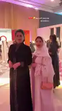 This stylish scene captures two women posing together at a glamorous indoor event, showcasing elegant attire and modern fashion. One woman wears a sleek black dress with intricate embroidery, while the other is dressed in a flowing white abaya adorned with delicate floral patterns and a matching hijab. They stand amidst a chic, minimalist setting with soft pink and purple lighting, surrounded by contemporary decor like oversized chess pieces and tall white columns. The atmosphere exudes sophistication and celebration, highlighting the essence of high-end fashion and cultural elegance. by @soja_design with Original Sound by soja_design