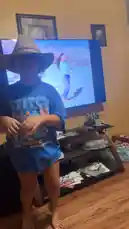 A young boy, identified as Tomas Guerrero's grandson, dances enthusiastically in his living room while wearing a large cowboy hat and a blue 'Stitch' t-shirt. He mimics the dance moves from a music video playing on the TV behind him, which features a woman singing. An adult male voice can be heard encouraging him to sing and dance along. by @tomasg3901 with Original Sound by tomasg3901