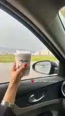 A person holds a coffee cup while driving through a scenic landscape. The view from inside the car shows a river, greenery, and distant buildings under a cloudy sky. The individual's hand, adorned with red nail polish and a watch, adds a personal touch to the tranquil journey. by @batoolmakeup1 with قهوتي المفضله من Vo❣️ by My Fav Coffee Of  Vo❣️SHahad