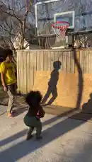 In a sunlit backyard, two young athletes engage in an energetic basketball session. One, shirtless with an afro, dribbles and shoots toward a hoop mounted on a wooden fence. His friend, wearing a yellow t-shirt and camouflage pants, watches closely before joining in. They take turns attempting dunks and shots, their long shadows stretching across the pavement. The scene captures the joy and dedication of youth sports, showcasing skill development and friendly competition in a casual outdoor setting. by @marcus.randall with Original Sound by marcus.randall