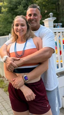 In this heartfelt scene, fitness influencer Mark The Shark embraces his partner outdoors after a workout. She wears a colorful sports tank top and maroon shorts, while he dons a white t-shirt and gray sweatpants. The couple shares a warm smile, highlighting their strong bond amidst a casual backyard setting with greenery and a white fence in the background. by @mts550 with original sound