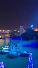 Experience the enchanting atmosphere of a city waterfront after dark. This captivating scene features glowing blue fountains dancing on the water, traditional round boats bobbing near a dock, and a vibrant city skyline illuminated by colorful lights in the background. by @guruofmarketing with Original Sound by guruofmarketing