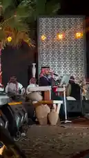 A captivating performance by a traditional Arabic music ensemble at an outdoor night event. The lead singer, dressed in a black thobe and red-and-white keffiyeh, passionately sings into a microphone while seated on a wooden chair. He is accompanied by musicians playing traditional instruments like the oud and percussion, all set against a beautifully lit stage with a geometric patterned backdrop, palm trees, and warm string lights. The entire scene exudes a festive and cultural atmosphere. by @jalsahevent with jalsahevent by Original Sound