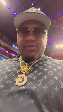 A man, identified as G-Mac Jazz, films himself at a Detroit Pistons game. He's wearing a Louis Vuitton shirt, a large gold chain with a 'G' pendant, and a black cap. He speaks to the camera before taking a drink from a white cup. The background shows the packed basketball arena and a large scoreboard. by @nbapistons with Original Sound by nbapistons