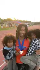 A joyful track and field athlete, wearing his medal, poses proudly with two young children on a running track at sunset. The moment captures the athlete's success and his connection with the next generation. by @marcus.randall with Original Sound by marcus.randall