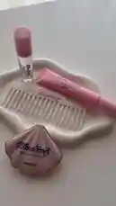 A close-up showcase of Blushing Beauty products, including a lip gloss, highlighter stick, and a decorative comb, all arranged neatly on a white dish. This aesthetic display highlights the brand's signature pink theme and product design. by @bx301a with 🪼🥂🏵️🌴 by Contains: The Realist - Soul Wun