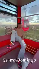 A man, identified as Delly, enjoys a scenic ride inside a red cable car in Santo Domingo. He sits relaxed on a bench, looking out at the cityscape and greenery below as the gondola moves along its track. The text 'Santo Domingo' is overlaid on the screen, confirming the location. by @delwinbazilme with La Forma en Que Me Miras by Super Yei, Sammy Baby