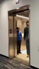A Boston Celtics player exits an elevator and walks confidently down a team corridor lined with branding. He wears a denim jacket over a white collared shirt and dark jeans, followed by media personnel with cameras. The hallway features prominent 'Boston Bruins' and 'JetBlue' sponsor logos, leading towards a tunnel entrance where more people gather. by @celtics with Original Sound by celtics