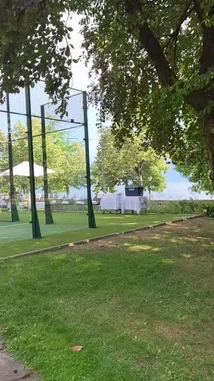 A serene lakeside tennis court complex comes to life with a perfectly arranged picnic setup. The camera glides across lush green courts, past elegant white-clothed tables laden with food, and onto modern lounge chairs, all set against the backdrop of a tranquil lake and a scenic promenade. This idyllic setting is the perfect destination for a relaxing day of sport and leisure. by @two with أحلى رسمه by فضل شاكر