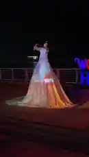 A performer plays the violin under the night sky, wearing a voluminous white gown. The fabric of her dress is illuminated with a vibrant, moving light projection, creating a stunning visual effect. The scene is set outdoors near a body of water, with a glowing sign visible in the background. by @home_garden3 with Original Sound by home_garden3