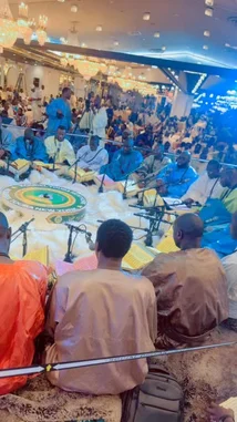 abdoufall205962 님이 만든 Original Sound 포함 @abdoufall205962 님이 만든 A large congregation gathers inside a grand mosque for a spiritual event led by Abdou Fall. Men dressed in traditional attire sit on prayer mats, attentively listening as he recites verses from the Quran. The ornate hall, illuminated by crystal chandeliers and vibrant blue stage lights, creates a solemn and reverent atmosphere.