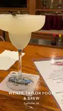 오리지널 사운드 포함 @brattyluhbaby 님이 만든 This scene captures a refreshing margarita served at Wenzil Taylor Food & Brew in Spillville, Iowa. The drink, garnished with a salt rim and black straw, sits elegantly on a wooden bar table next to a menu and a Daytona 500 coaster. The warm, inviting ambiance suggests a casual dining experience perfect for enjoying craft cocktails and local cuisine.