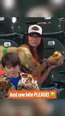 A woman enjoys a hot dog at a baseball game while her golden retriever sits beside her, begging intensely for just one bite. The dog's pleading eyes and eager behavior create a humorous and heartwarming moment. by @mlb with Original Sound by mlb