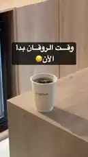 A close-up shot captures a white paper cup from Sequence Coffee resting on a modern wooden counter. Steam gently rises from the dark coffee inside, creating a serene and inviting atmosphere. In the background, a glass-fronted refrigerator adds to the contemporary setting. A child's voice can be heard saying 'Bida wakt rokan,' adding a candid, everyday moment to the scene. by @sequence-coffee with الحين بدأ وقت روقان 😂 by a