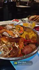 A close-up view of a massive seafood boil served in a large metal bowl, featuring crab legs, crawfish, sausage, corn on the cob, and potatoes. The dish is presented on a table in a bustling restaurant setting, with diners visible in the background. A text overlay displays the song 'What I Want' by Morgan Wallen and Tate McRae, suggesting a lively dining atmosphere. by @tomasg3901 with What I Want by Morgan Wallen, Tate McRae