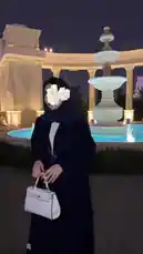 A woman in a chic black abaya and hijab poses gracefully in front of a beautifully lit fountain at night. She holds a white handbag and adjusts her outfit, showcasing her fashionable look against the backdrop of classical architecture. by @soja_design with original sound